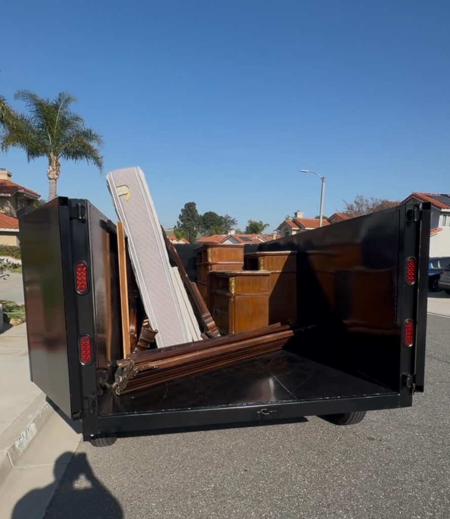 A black trailer loaded with furniture, including a mattress, wooden cabinets, and wooden frames.