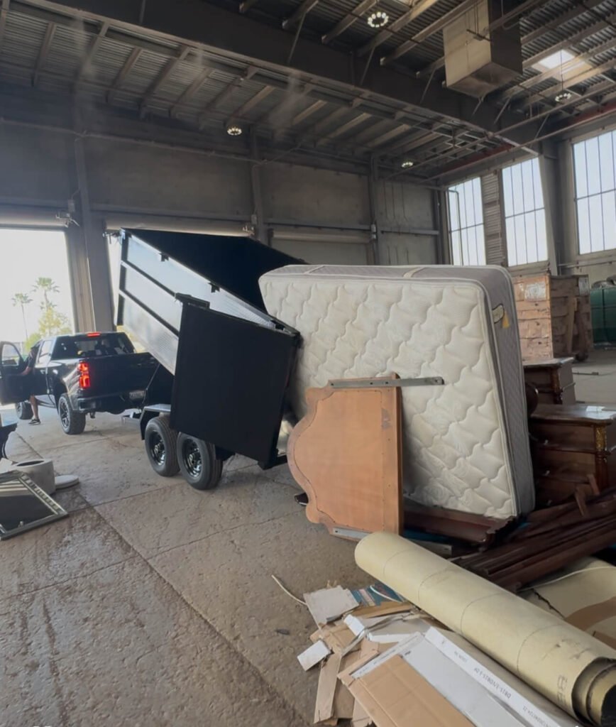 Truck unloading a mattress and furniture into a trailer in an industrial area.